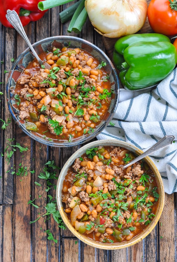Overhead image of two bowls of slow cooker pork and beans with ground beef surrounded by vegetables