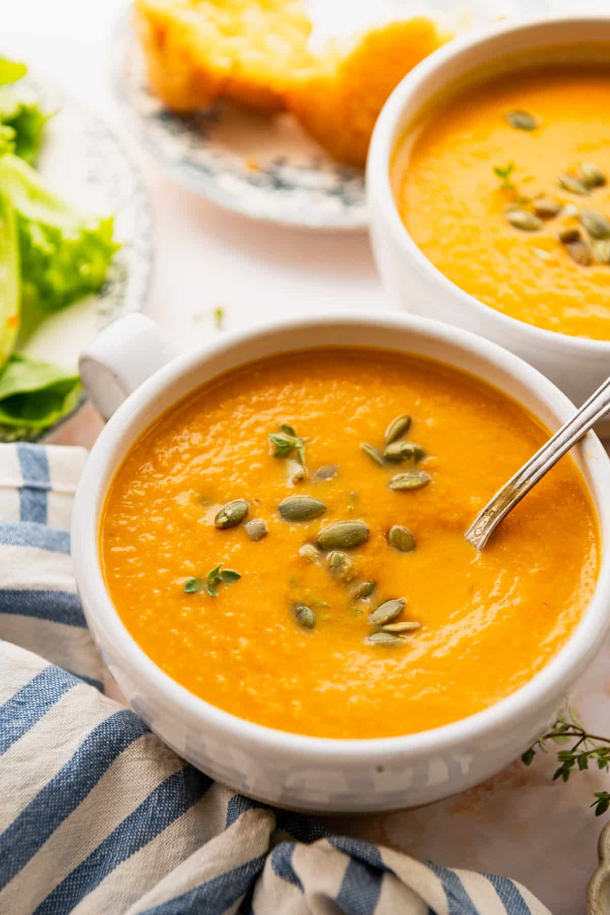 Side shot of two bowls of healthy and easy pumpkin soup served in white bowls with pumpkin seeds on top for a garnish.