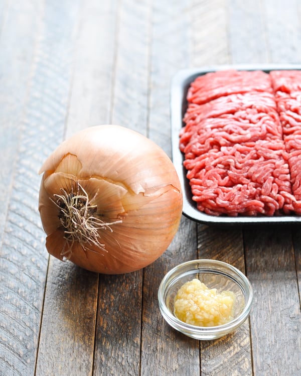 A large yellow onion, minced garlic, and ground beef laid out on a wooden table top - all ingredients to make Chili con Carne