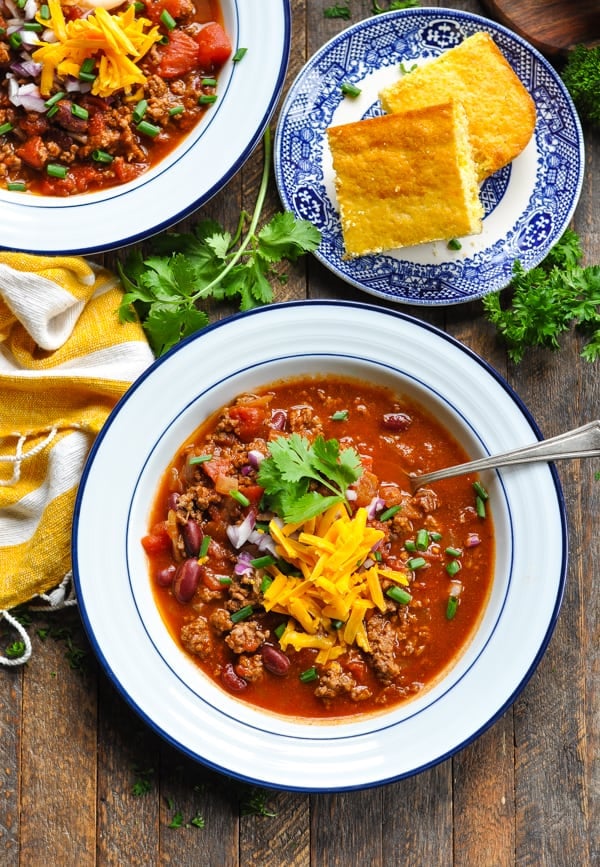 An overhead shot of two bowls of homemade chili con carne, both garnished with shredded cheese, fresh cilantro, and cornbread.