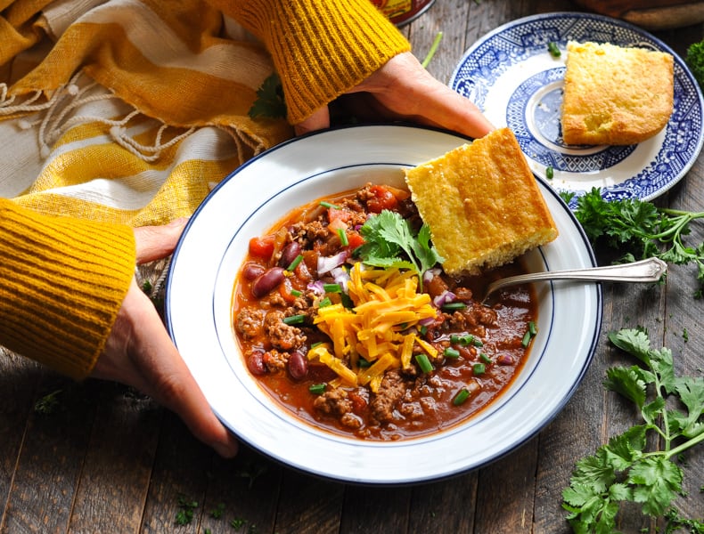 A woman's hands hold a bowl of Chili con Carne served with a side of cornbread. She's wearing a long sleeved yellow sweater.