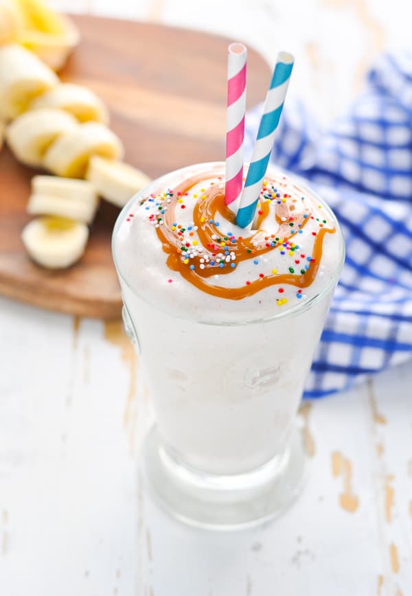 Healthy banana smoothie in a glass with pink and blue straws and caramel sauce on top