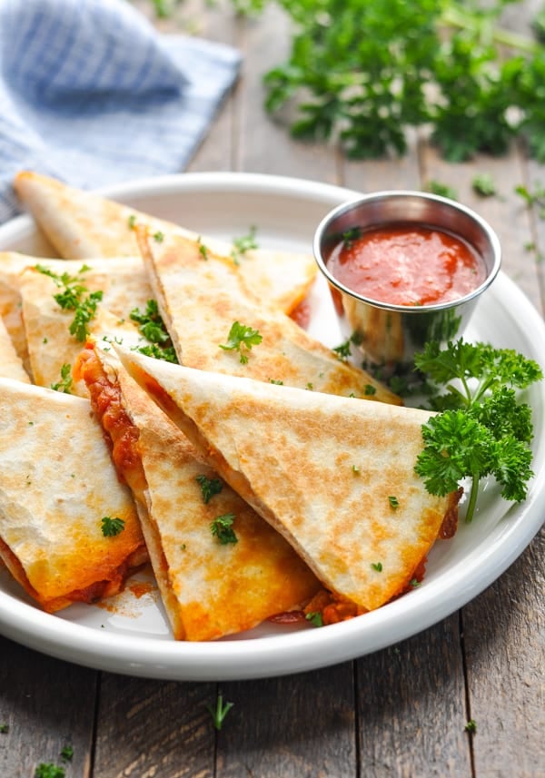Pepperoni pizza quesadilla triangles stacked on a white plate, served with a side of marinara dipping sauce.