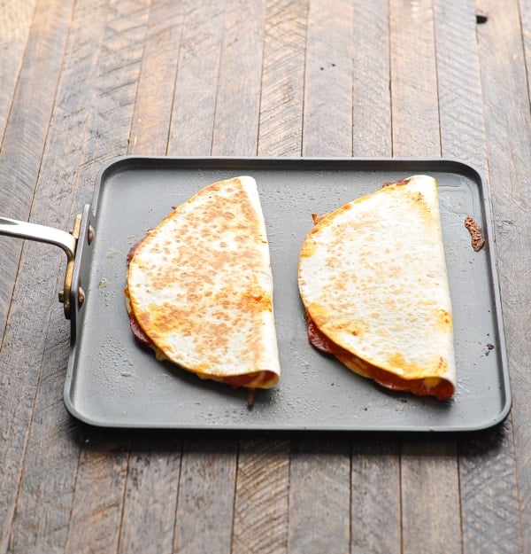 Two crispy pizza quesadillas sit on a skillet pan set on a wooden table.