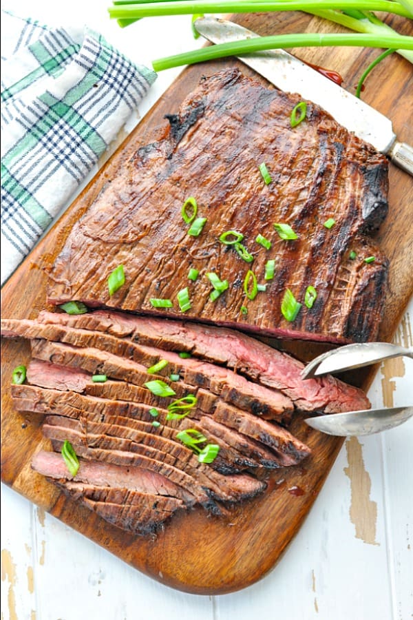 Sliced flank steak on a cutting board with knife and tongs