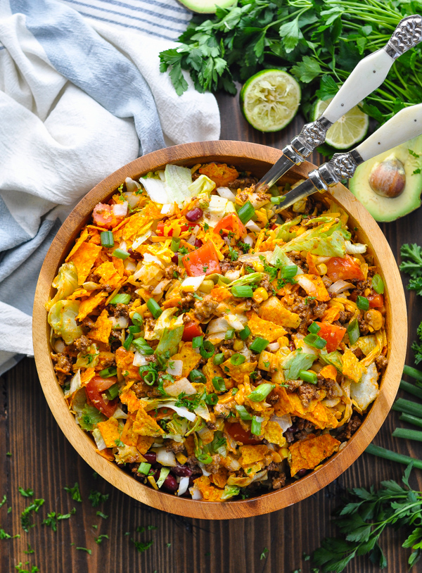 Overhead shot of taco salad in a wooden bowl surrounded by avocado and fresh herbs