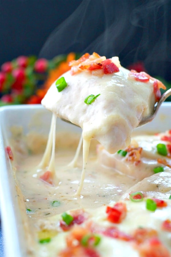 A person uses a serving spoon to lift a single chicken breast from a pan of creamy chicken bacon ranch alfredo. The chicken is smothered in a creamy sauce and topped with bacon pits and sliced green onions.