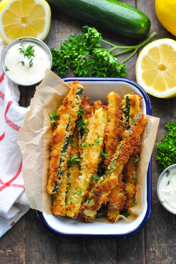 Overhead shot of dish of zucchini fries surrounded by herbs lemon and dip