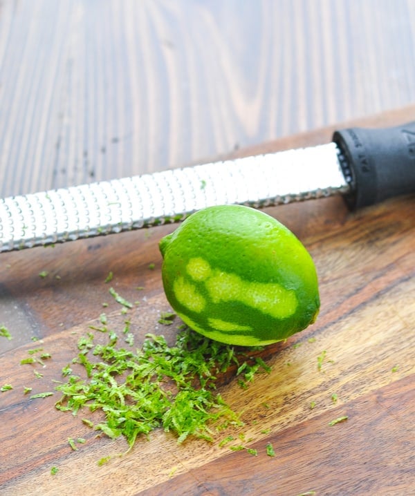 A partially zested lime rests on a cutting board next to a microplane grater.