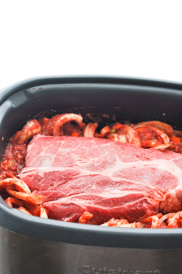 A raw chuck roast sits atop of a tomato sauce and onion mixture in a large slow cooker.