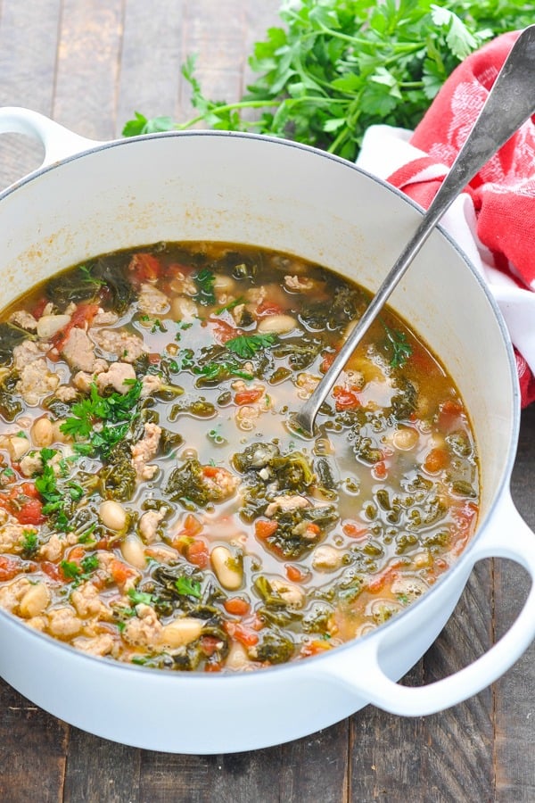 Ladle in a white Dutch oven full of Tuscan White Bean Soup with Sausage and Kale