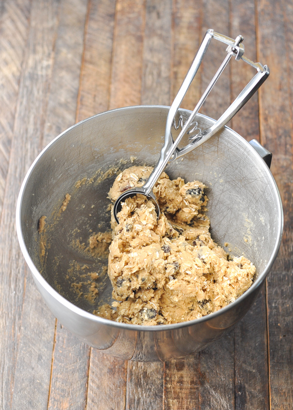 Raw oatmeal raisin cookie dough in a stand mixer bowl with a spring-loaded scooper scooping out a ball of cookie dough.