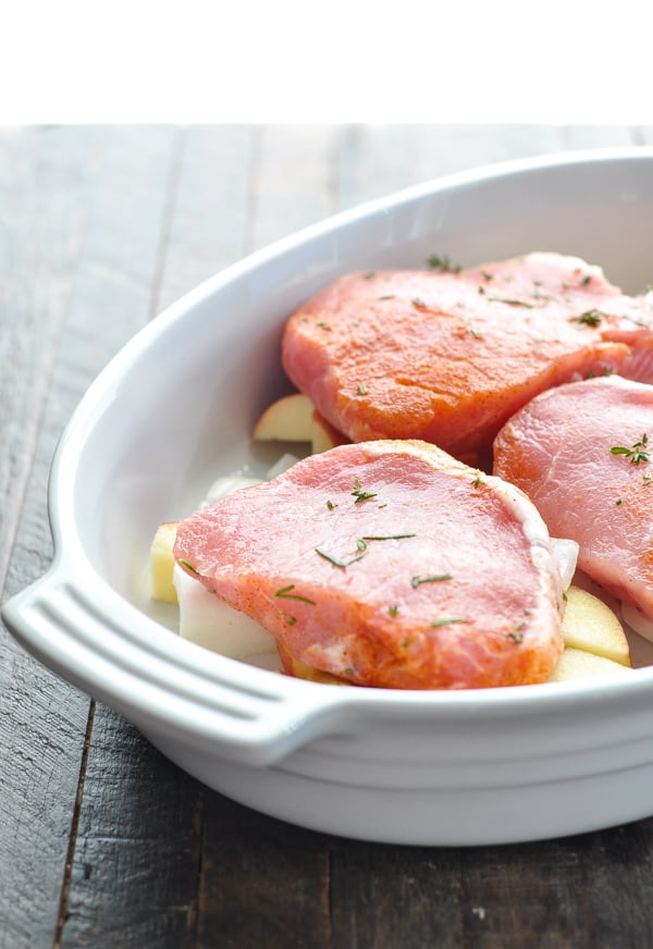 Raw pork chops in a white baking dish on top of apples and onions
