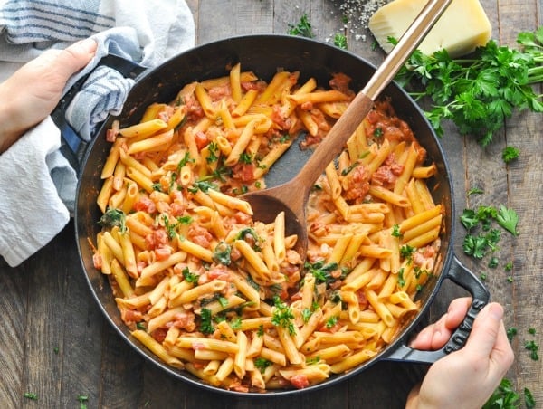 A woman serves a pot of spinach and tomato pasta on a wooden tabletop. Fresh parsley and a block of parmesan cheese sit on the table beside the cast iron skillet.