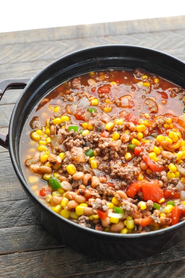 A large cast iron pot is filled with chili ingredients - cooked ground beef and sausage mixed with black eyed peas, corn, bell peppers, and diced tomatoes in a liquidy broth.