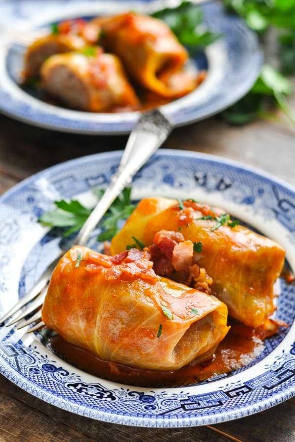 Two stuffed cabbage rolls sit on a plate, covered in a hearty tomato sauce and garnished with bacon and parsley.