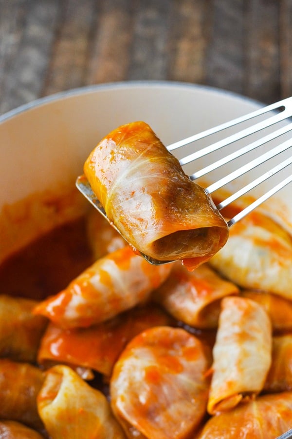 A single cooked stuffed cabbage roll covered in tomato sauce sits on the paddle of a spatula. A whole pot of fresh cooked cabbage rolls is in the background.