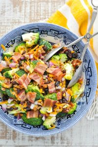 Close overhead shot of a bowl of broccoli salad with bacon and silver serving tongs