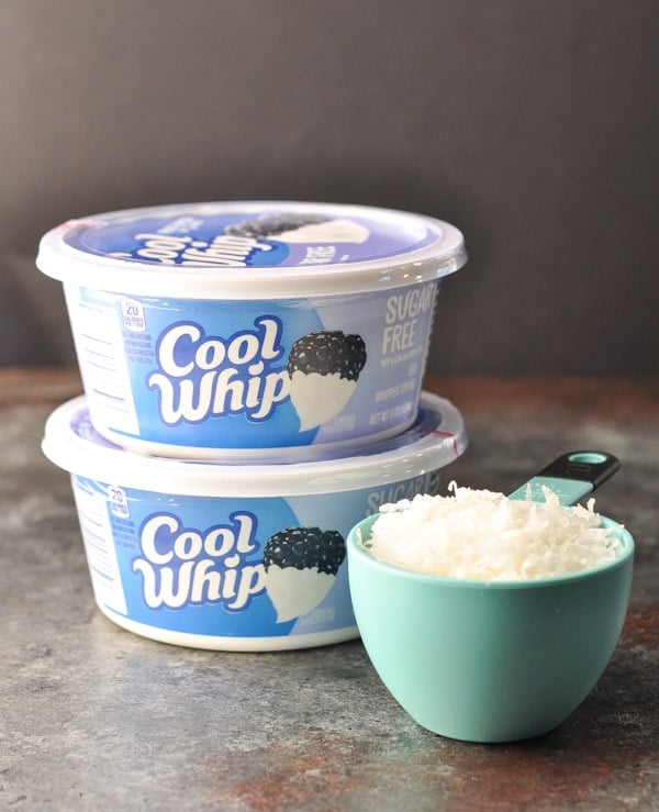 Two containers of sugar free cool whip stacked on top of each other, next to a measuring cup filled with sweetened coconut flakes.