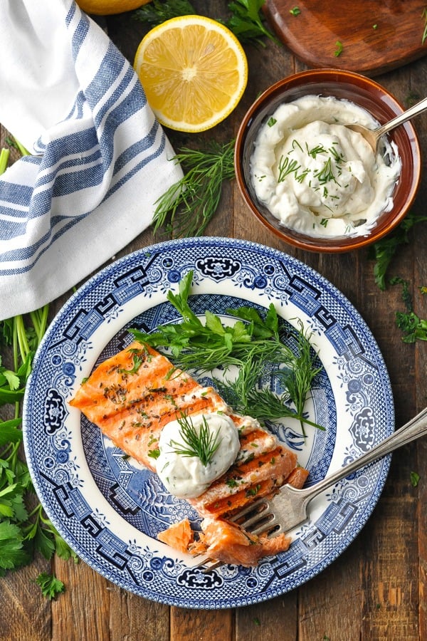 Grilled salmon with dill sauce served on a blue plate, next to a bowl of creamy homemade dill ypgurt sauce.
