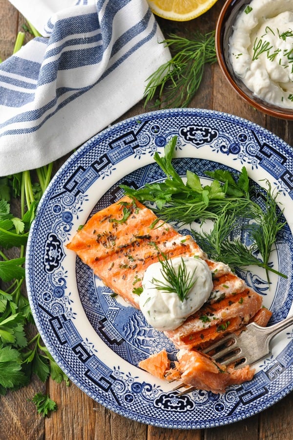 A grilled salmon fillet topped with a creamy dill yogurt sauce, served on a plate that's garnished with fresh dill and parsley.