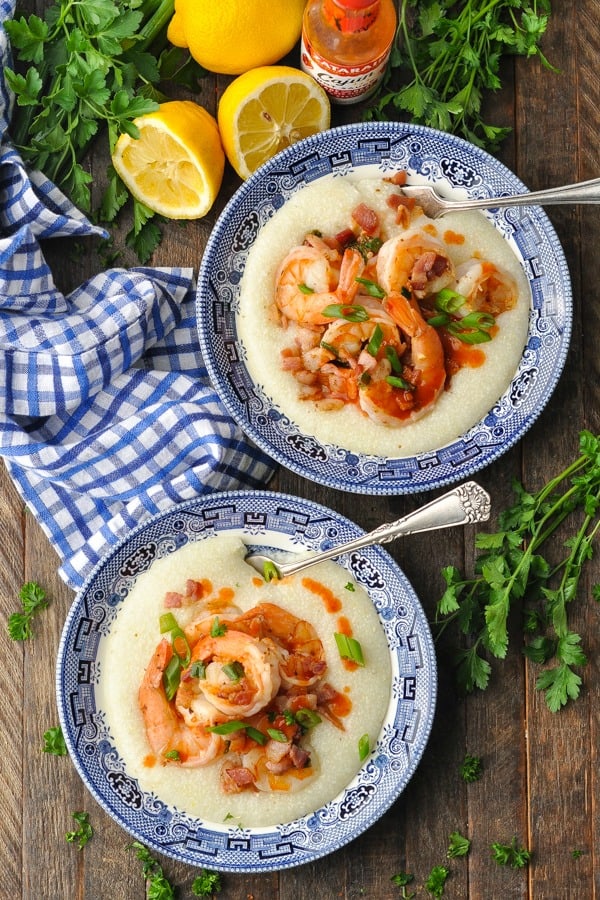 Overhead shot of shrimp and grits served in bowls and surrounded by hot sauce, lemon and fresh herbs