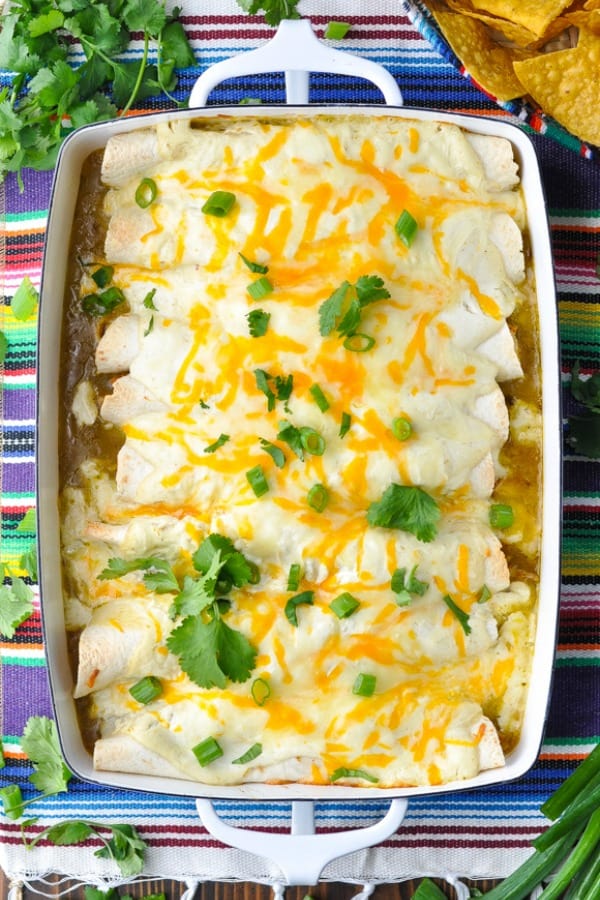 Overhead shot of a pan of sour cream chicken enchiladas with green sauce in a white dish on a striped tablecloth