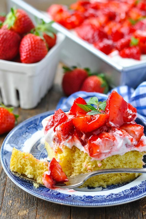 A close up image of a single slice of Strawberry Shortcake Cake served on a dessert plate. A fork rests on the plate, holding a small bite of cake.