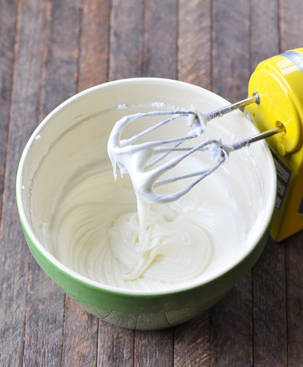 Whisks of a hand mixer rest on the side of a mixing bowl, filled with a smooth cream cheese frosting.