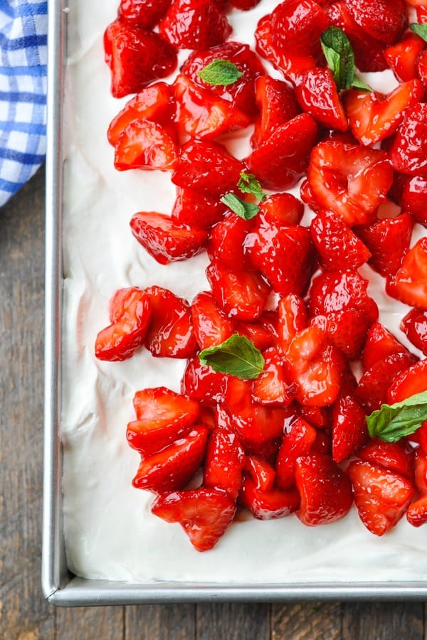A close up overhead image of glazed strawberries spread out over a Strawberry Shortcake Cake iced with a whipped topping and fresh mint leaves garnish.
