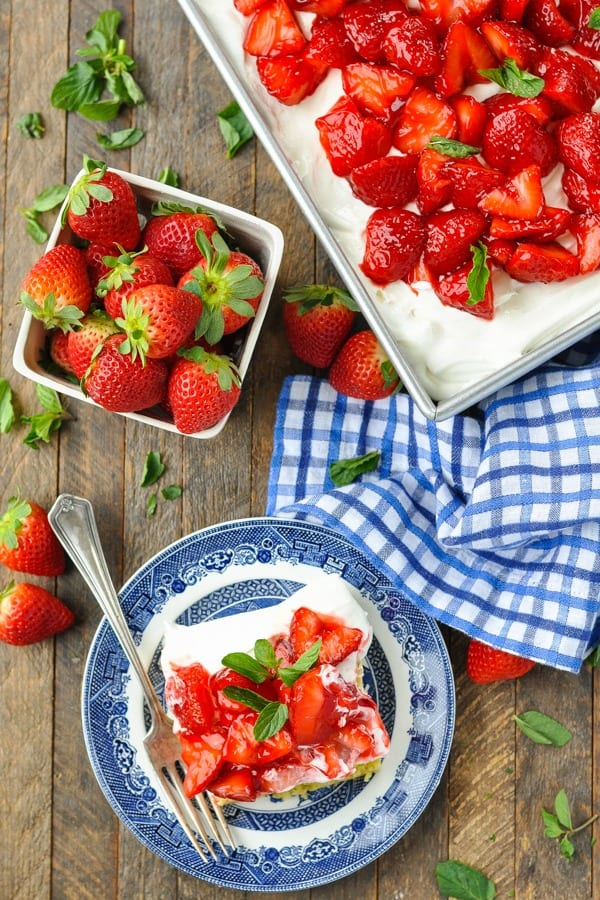 An overhead image of a dessert table filled with a whole cake pan of Strawberry Shortcake Cake, a single sliced served on a small blue and white plate, and a bowl of fresh strawberries.