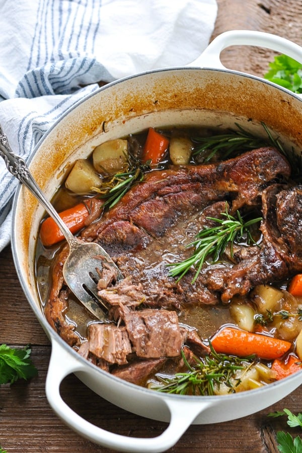 Side shot of a dutch oven pot roast with carrots and potatoes and fresh herbs in a white pot