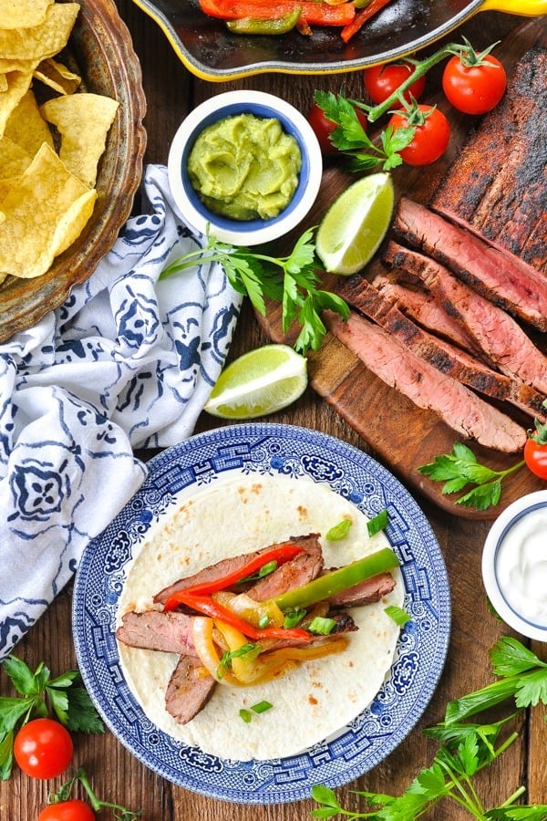 Overhead shot of a steak fajitas recipe on a table surrounded by limes chips and guacamole