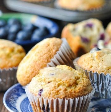 Front shot of three easy blueberry muffins on a blue and white serving plate with more muffins and fresh berries in the background
