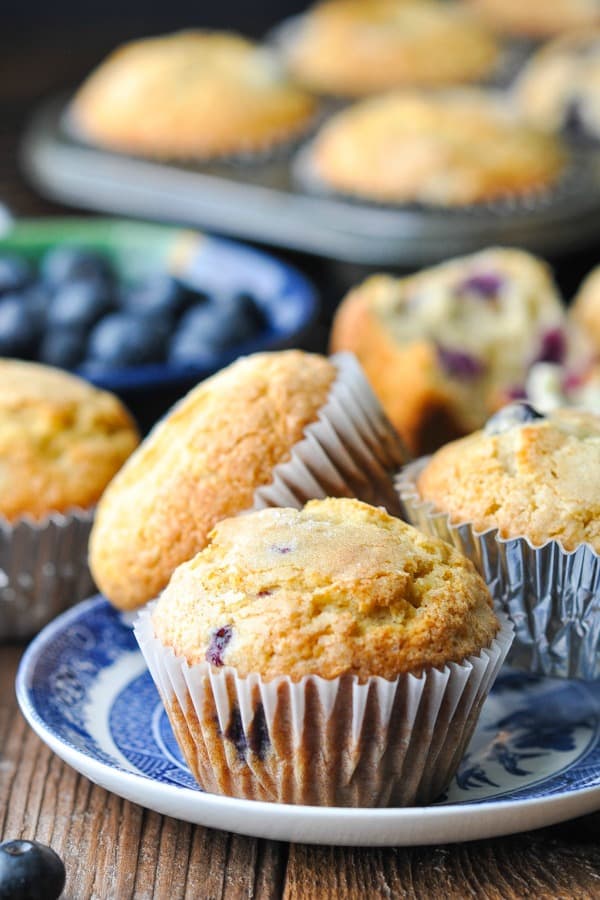 Front shot of three easy blueberry muffins on a blue and white serving plate with more muffins and fresh berries in the background