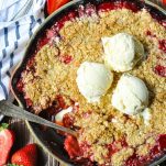Spoon digging into a skillet of warm strawberry crisp with vanilla ice cream on top