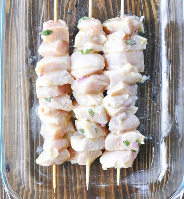 Marinated chicken souvlaki on wooden skewers in a glass dish before cooking.