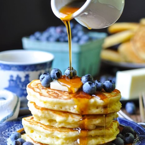 A plate piled high with blueberries and fluffy pancakes while syrup is poured over top