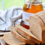 Front shot of a loaf of honey wheat bread sliced on a wooden cutting board with a jar of honey in the background