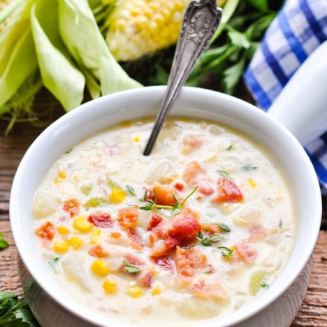 An easy corn chowder recipe served in a white bowl with bacon on top and fresh corn in the background