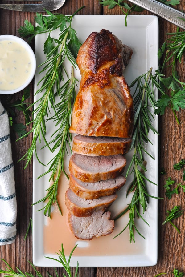 Long overhead shot of pork tenderloin sliced on a white serving platter with fresh rosemary and mustard sauce nearby