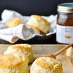 Front shot of a tray of buttermilk biscuits with honey and a basket in the background
