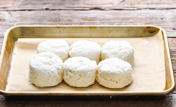 Six raw buttermilk biscuits lined up on a parchment paper-lined baking sheet, ready to go into the oven.