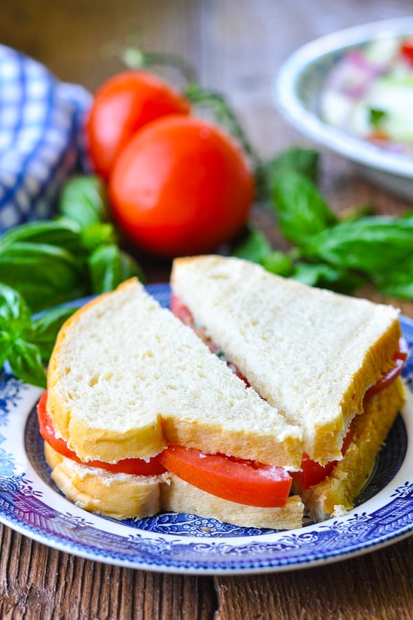 Front shot of a tomato sandwich on a blue and white plate with fresh basil and tomatoes in the background