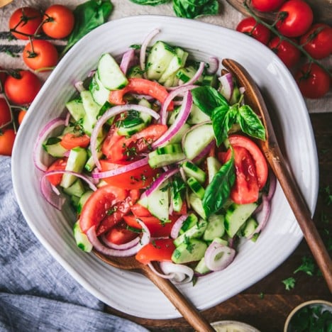 Square overhead featured image of a cucumber tomato onion salad in a white bowl with wooden serving spoons
