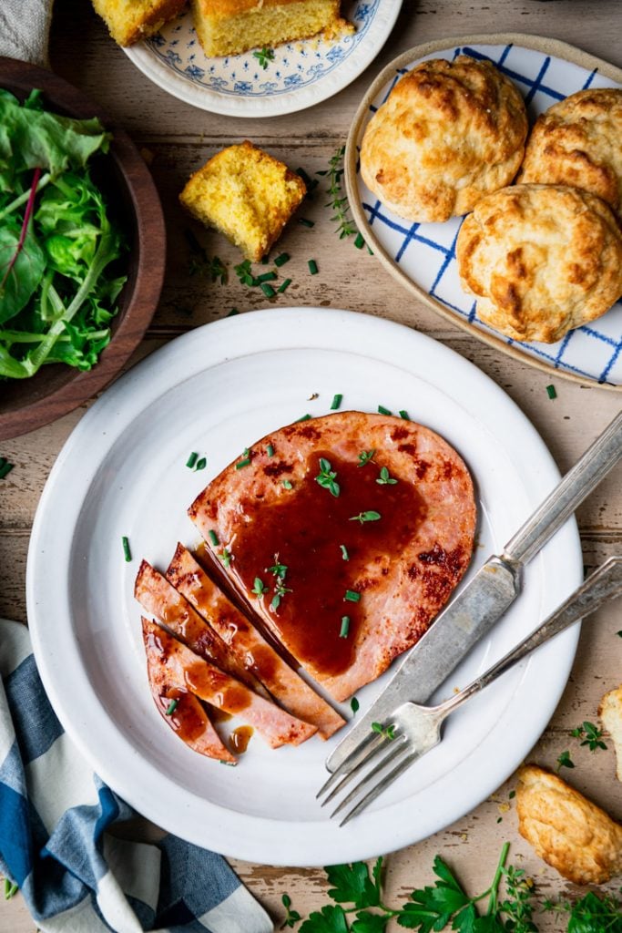 Glazed ham steak recipe on a white plate with biscuits and salad on the side