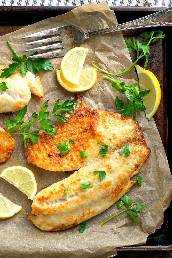 Overhead shot of baked tilapia fillet surrounded by fresh parsley and lemon