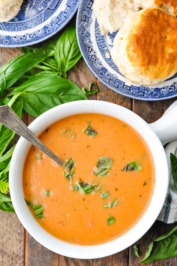 Overhead image of a bowl of roasted tomato soup on a wooden table surrounded by fresh basil