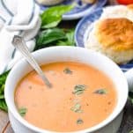 Front shot of a bowl of roasted tomato soup on a table with basil and biscuits in the background