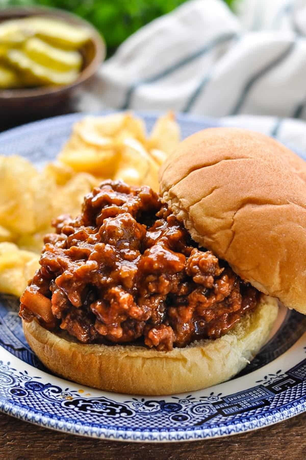 Side shot of an old fashioned sloppy joes recipe served on a sandwich bun with pickles and potato chips.